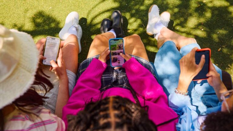An overhead shot of three young girls wearing colourful clothes using smartphones sitting on a bench in a park.